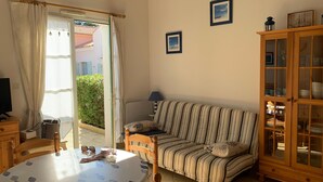 A living room with a striped sofa, a wooden cabinet with glass doors, a dining table with a fruit pattern tablecloth, and a television on a wooden stand. - House near beach, pets welcome - 4 guests (Saint-Hilaire-de-Riez)