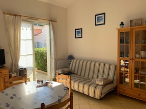 A living room with a striped sofa, a wooden cabinet with glass doors, a dining table with a fruit pattern tablecloth, and a television on a wooden stand. - House near beach, pets welcome - 4 guests (Saint-Hilaire-de-Riez)