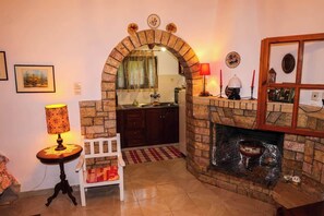 Ein Wohnzimmer mit Kamin, einem weißen Sessel, einem Tisch mit Lampe und einer durch einen Bogen sichtbaren Küche.
