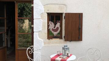 Un bâtiment rustique avec une porte et des volets en bois, une décoration représentant un coq, et deux chaises en métal blanc disposées autour d’une table préparée pour deux personnes.