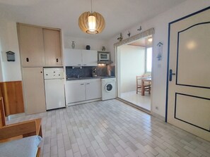 A compact kitchen with white cabinets, a washing machine, a microwave, and a dining area with a table and chairs. - T2 Cabin Apartment - 35m² - Saint-Pierre-La-Mer (Fleury-Saint-Pierre-la-Mer)