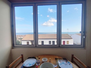 A dining area with a table set for two, a window with a view of buildings and the sea. - T2 Cabin Apartment - 35m² - Saint-Pierre-La-Mer (Fleury-Saint-Pierre-la-Mer)