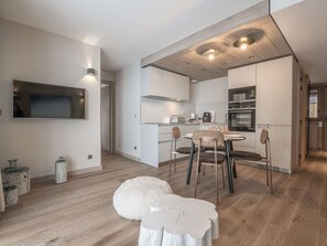 Eine moderne Küche mit weißen Schränken, eine Essecke mit rundem Tisch und Stühlen, sowie ein an der Wand befestigter Fernseher.