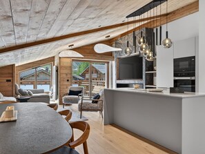 Ein modernes Interieur mit Holzbalkendecke, einem Essbereich mit rundem Tisch und Stühlen, einer Küche mit Einbauofen und Mikrowelle sowie einem großen Fenster mit Blick auf eine Berghütte.