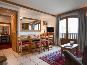 A cozy living room with a wooden dining table, chairs, a television, and a large window with curtains. - Cozy apartment in the center - 4 guests (Courchevel)