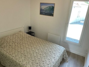 A bedroom with a bed, a nightstand, a wall-mounted lamp, and a window with a view of a building and a street. - House with parking and garden - 4 guests (Capbreton)