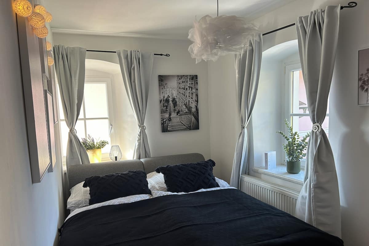 Una cama con ropa de cama blanco y negro, cortinas grises, una lámpara de pared e imagen en un marco.