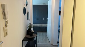 Un couloir mène à une pièce dont le sol est en bois, où se trouve une petite table avec une plante et un téléphone fixé au mur.