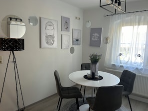 Un área de comedor moderna con una mesa redonda, cuatro sillas, una lámpara de pie y decoración en la pared que incluye un retrato y elementos con motivos marinos.