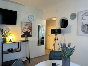 Una sala de estar moderna con una televisión de pantalla plana, un reloj, un retrato enmarcado, una lámpara de pie y una zona de comedor con mesa y sillas.