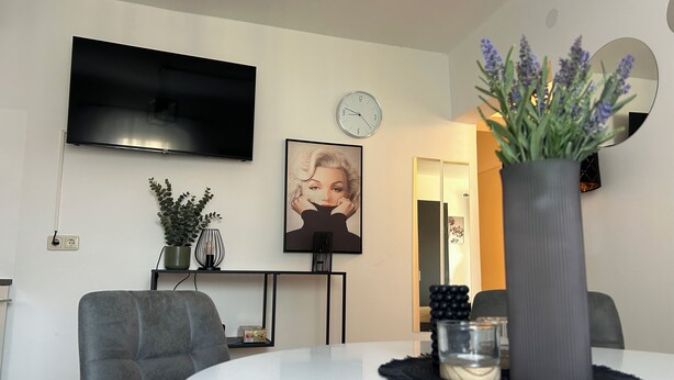 Un salon moderne avec un téléviseur à écran plat, une horloge, un tableau encadré, une plante en pot et une table de salle à manger ronde décorée de bougies.