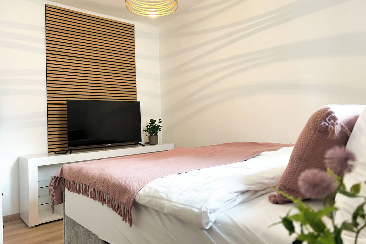 Un dormitorio con cama, televisor sobre un mueble, estante montado en la pared y lámpara en el techo.