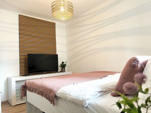Un dormitorio con cama, televisor sobre un mueble, estante montado en la pared y lámpara en el techo.