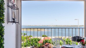 Un balcon donnant sur la mer, une table à manger dressée avec des assiettes et des couverts, et une cage à oiseaux accrochée au mur.