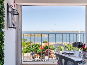 Ein Balkon mit Blick auf das Meer, ein Esstisch gedeckt mit Tellern und Besteck sowie ein Vogelkäfig an der Wand.