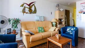 Un salon comprenant un canapé, un fauteuil bleu, une table basse en bois, un ventilateur, une plante en pot et un élément décoratif mural.