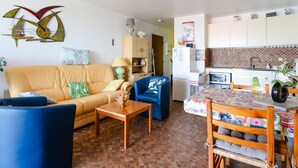 Un salon comprenant un canapé, une table basse, un fauteuil bleu et un coin repas avec une table et des chaises. Il y a également une cuisine équipée de placards et d’un réfrigérateur.
