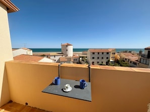 A balcony with a table, two cups, and a view of a coastal town with buildings and a tower. - T2 Apartment sea view, parking - 4 guests (Fleury-Saint-Pierre-la-Mer)