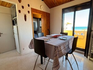 A dining area with a table set for two, a clock on the wall, and a view of the sea through a glass door. - T2 Apartment sea view, parking - 4 guests (Fleury-Saint-Pierre-la-Mer)