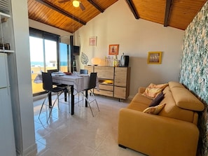 A living room with a sofa, a dining table with chairs, a wooden cabinet, and a ceiling fan. - T2 Apartment sea view, parking - 4 guests (Fleury-Saint-Pierre-la-Mer)