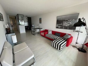 Ein modernes Wohnzimmer mit einem roten Sofa, einem schwarz-weiß gestreiften Hocker, einem Esstisch mit Stühlen, einer Küchenzeile und einem großen schwarz-weißen Stadtbild an der Wand.