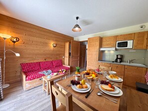 Ein gemütliches Zimmer mit einer Holzcouch, einem Esstisch mit Tellern, Gläsern und Besteck sowie einer Küchenzeile mit einer Mikrowelle.