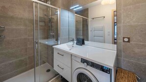 Une salle de bain moderne avec une douche à parois de verre, un lavabo blanc, un grand miroir et un lave-linge encastré.
