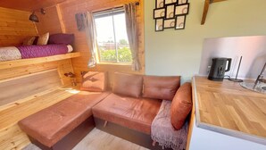 Un interior acogedor de cabaña con techo de madera, un sofá, un ventanal con cortinas, un marco de fotos colgado en la pared y una tetera sobre la encimera.