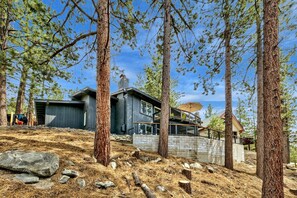 A modern cabin with a stone foundation, surrounded by tall pine trees, and a wooden deck with a glass railing. - 4 Bedrooms, Lakeviews, Hot Tub, Pool Table! (South Lake Tahoe)