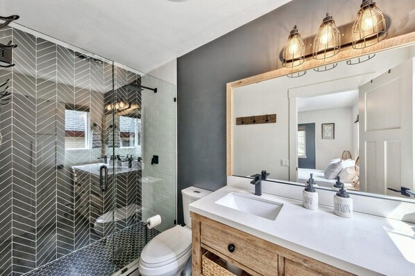 Un baño moderno con cabina de ducha de vidrio, un lavamanos blanco con dispensadores de jabón y loción, y un espejo grande con marco de madera.