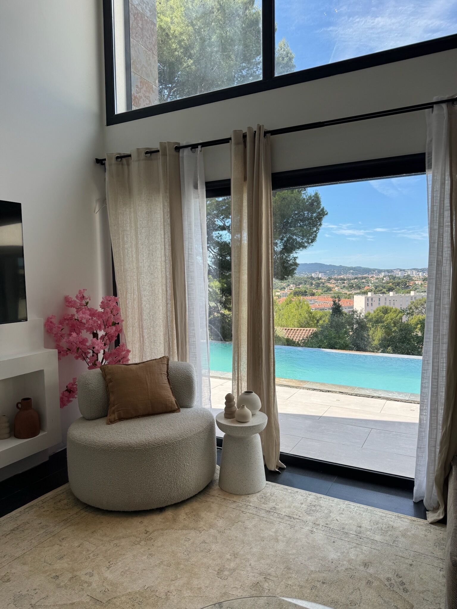 Un salon moderne avec un espace salon circulaire, une petite table d’appoint, un téléviseur fixé au mur et une grande fenêtre donnant sur une piscine et un paysage verdoyant.