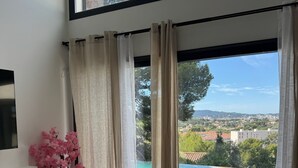 A modern living room with a round seating area, a small side table, a TV mounted on the wall, and a large window with a view of a pool and landscape. - TOULON ARCHITECT-DESIGNED VILLA (Toulon)