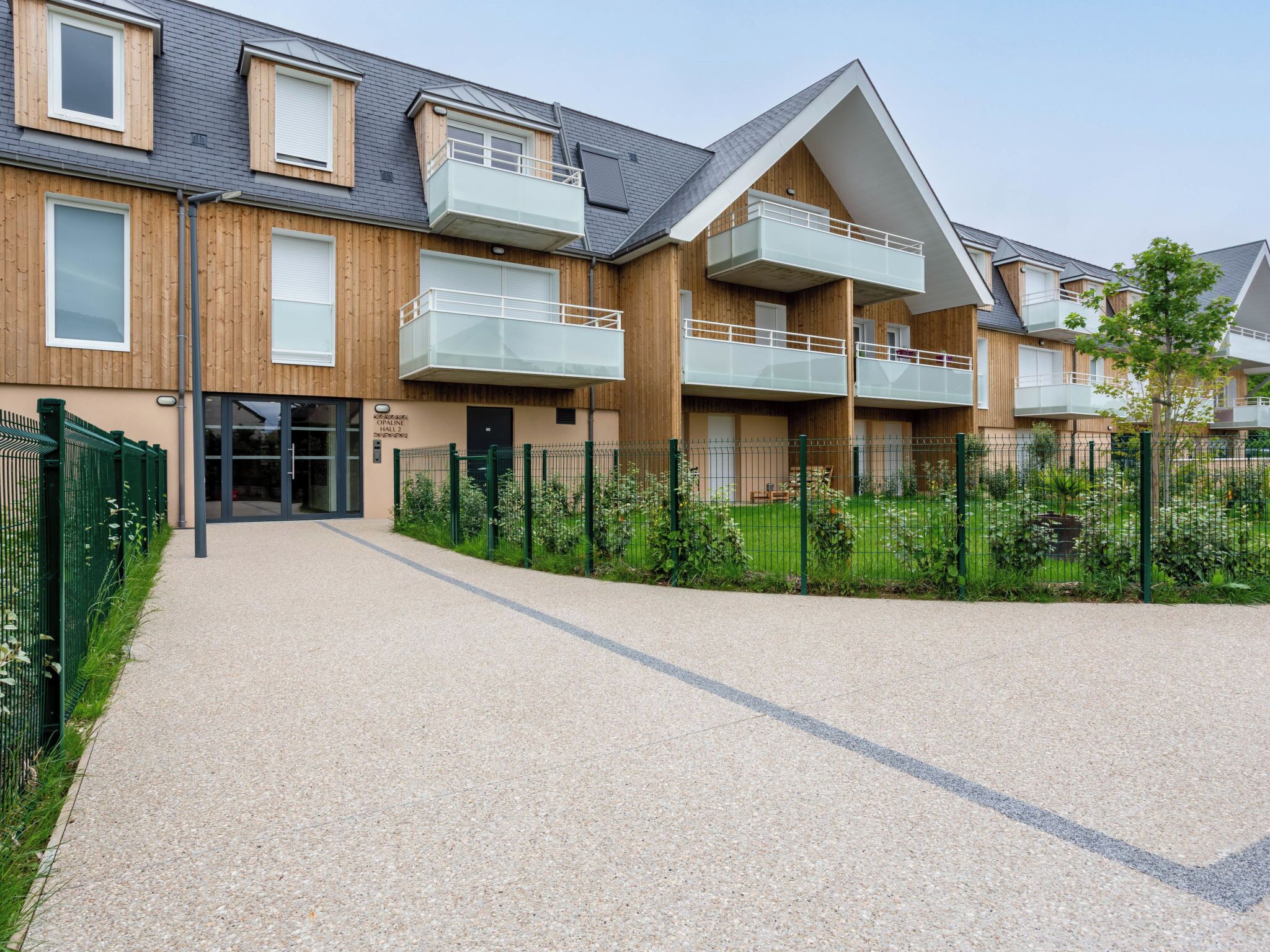 A modern residential building with wooden paneling, multiple balconies, and a gravel driveway.