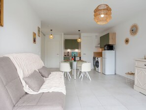Un salon moderne comprenant un canapé, une table à manger avec des chaises, un coin cuisine équipé d’appareils électroménagers et un luminaire suspendu.