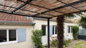 Een overdekt terras met een tafel en stoelen, een palmboom en een huis met een betegeld dak en ramen.