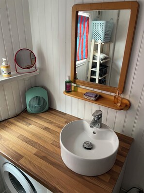 A bathroom with a wooden vanity, a white sink, a mirror, a shelf with toiletries, a fan, and a washing machine. - Charming house facing the Atlantic, garden and pergola 2 min from beaches (Fouras)