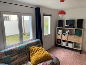 A room with a sofa, a yellow pillow, a TV on a stand, a bookshelf, and a window with a curtain. - Charming house facing the Atlantic, garden and pergola 2 min from beaches (Fouras)