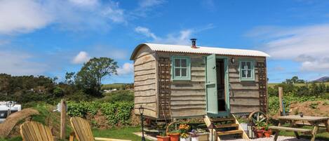A wooden cabin with a door, windows, and a wheelhouse. There are wooden chairs and a picnic table outside.