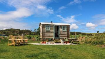 Una pequeña cabaña de madera con la puerta abierta, rodeada de un césped con mobiliario de exterior.