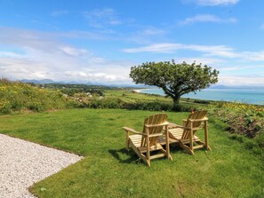 A grassy area with two wooden chairs facing a tree and a view of the sea. - Cwt Bugail (Pwllheli)
