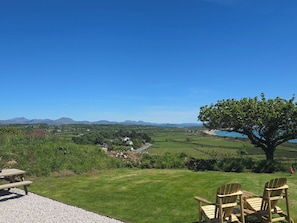 Un espacio al aire libre con césped, una mesa de picnic y dos sillas Adirondack, con vistas a un paisaje pintoresco con colinas y una masa de agua.