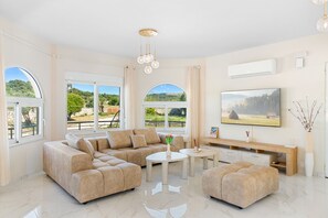 Ein geräumiges Wohnzimmer mit einem Ecksofa, einem Couchtisch, einem Fernseher und großen Fenstern mit Blick ins Freie.