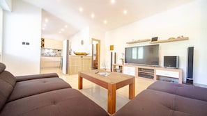 Ein modernes Wohnzimmer mit einer Sofagarnitur, einem Holztisch, einem Fernseher und einer Küche im Hintergrund.