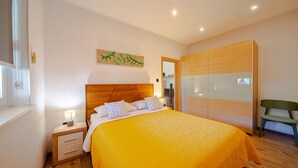 Un dormitorio con cabecera de madera, una cama con manta amarilla, una mesita de noche con lámpara, una silla verde y un armario con puertas espejadas.