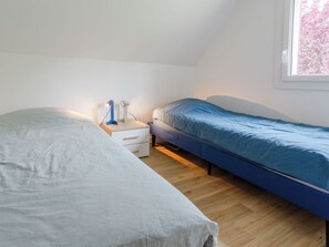 Ein Schlafzimmer mit zwei Betten, einem Nachttisch, einer Lampe und einem Fenster mit Blick auf Bäume.