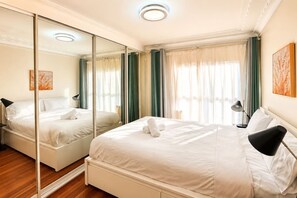 A hotel room with a large mirror, a bed with white linens, a bedside table with a lamp, and a window with curtains. - 3BR Mascot House | 3 Queen Beds | Walk to Station (Mascot)