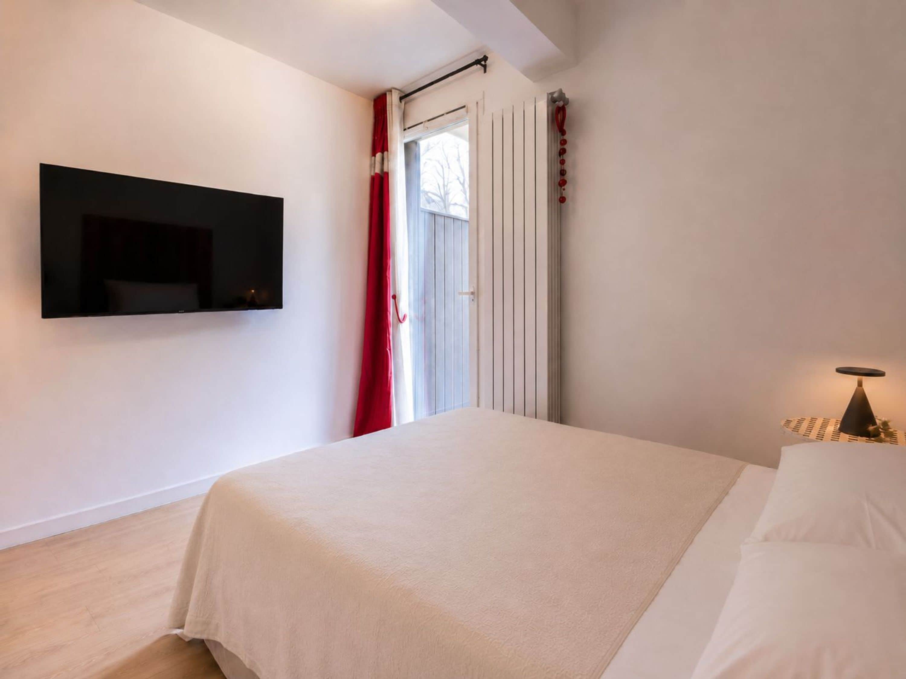Un dormitorio con cama, televisor montado en la pared, ventana con cortinas y una lámpara de noche.