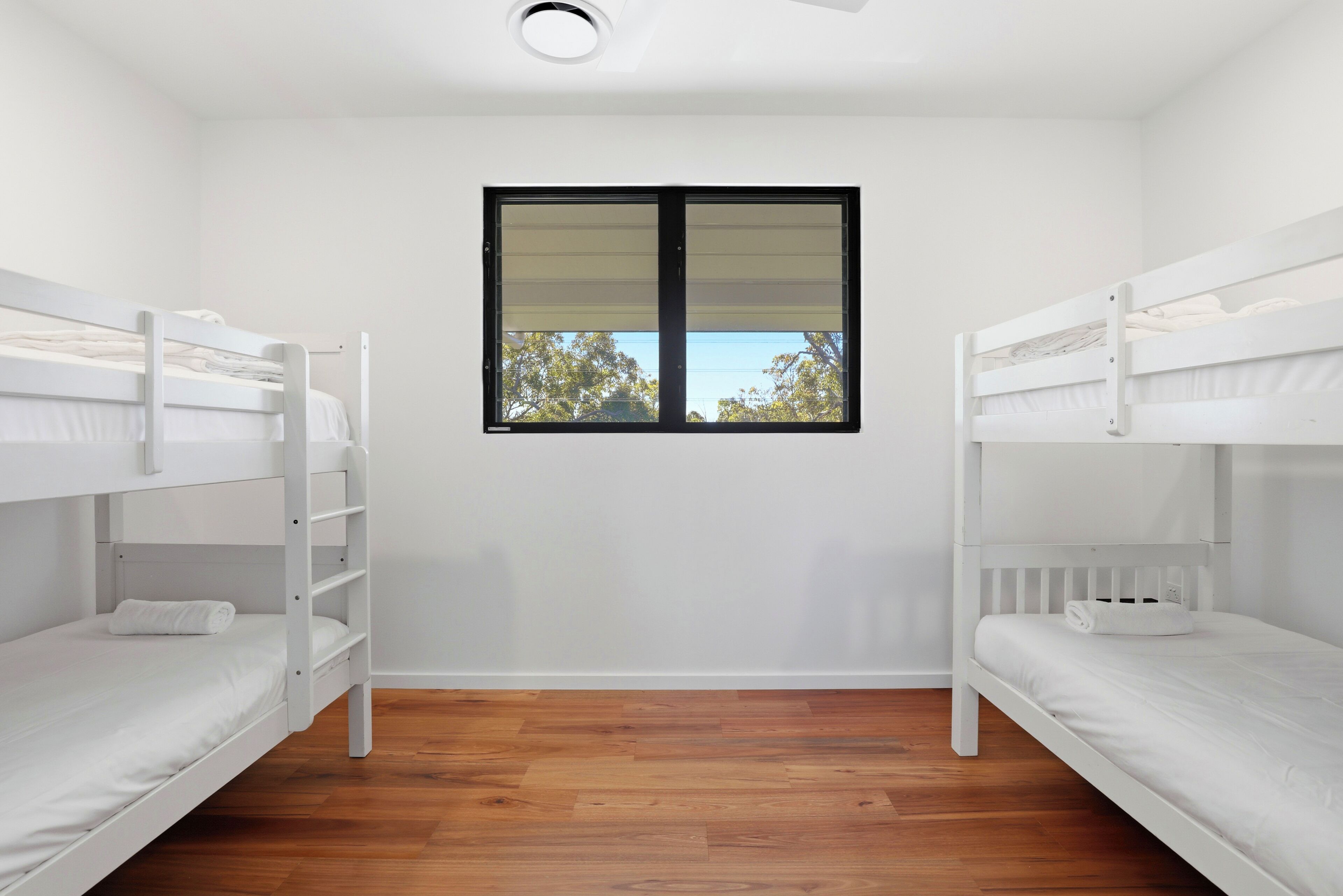 Une pièce avec deux lits superposés, une fenêtre avec des stores, un plancher en bois et un luminaire au plafond.