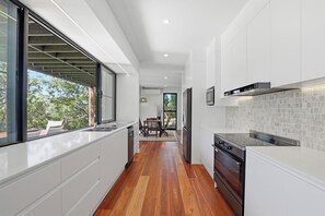 A modern kitchen with white cabinetry, a wooden floor, and a view through the window. - Tree House by the Sea | Modern Beach Home (Point Lookout)