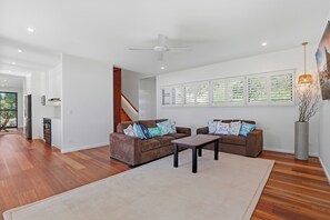 A modern living room with a brown sofa set, a coffee table, and a large window with blinds. - Tree House by the Sea | Modern Beach Home (Point Lookout)